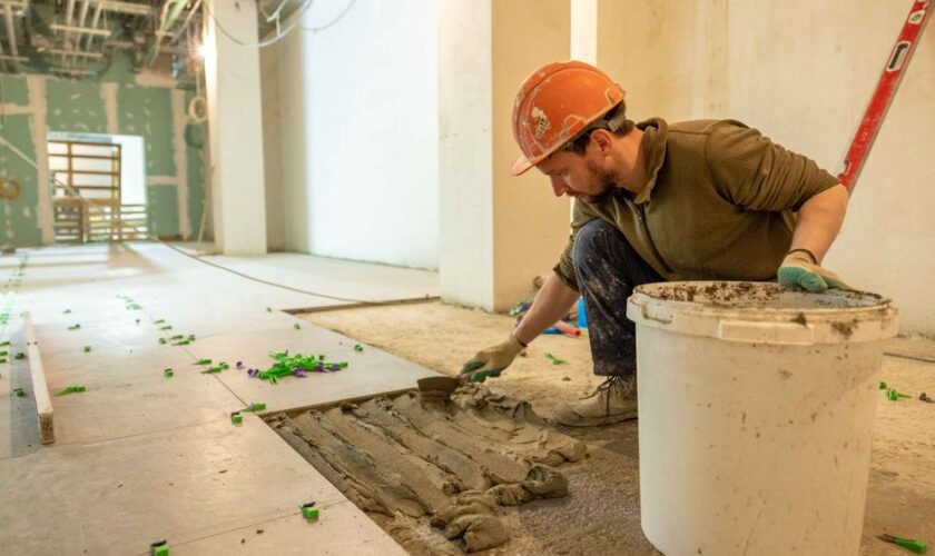A construction worker lays tiles in an indoor renovation project, wearing safety gear.