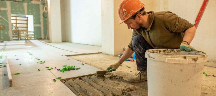 A construction worker lays tiles in an indoor renovation project, wearing safety gear.