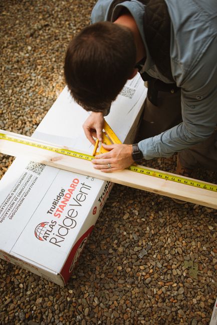 A construction worker measuring timber with a tape measure on a gravel worksite.