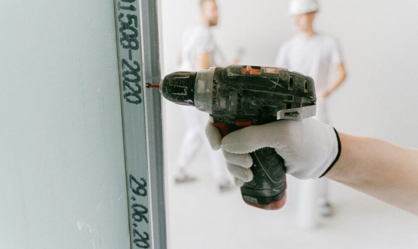 A construction worker uses a drill on drywall with blurred workers in the background.
