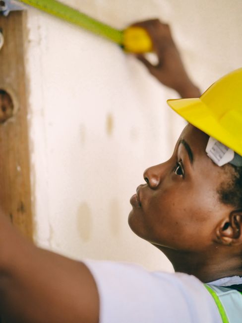 A construction worker using a measuring tape on a wall, wearing a yellow hardhat.
