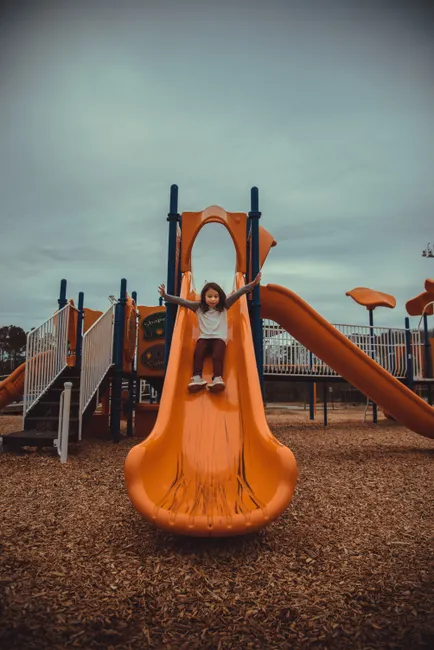 A joyful child sliding down an orange slide at an outdoor playground, expressing excitement.