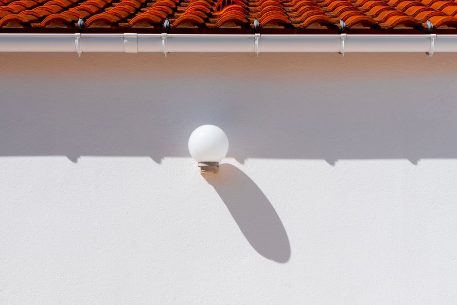 A minimalist outdoor setting showcasing a wall lamp casting a shadow beneath a red tiled roof.