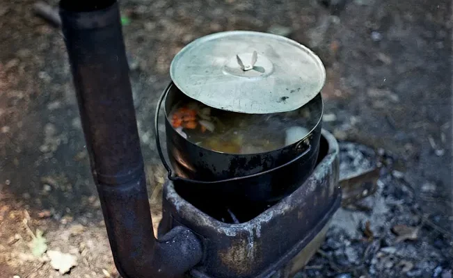 A rustic wood stove with a pot of food outdoors in a natural setting.