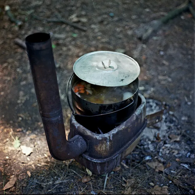 A rustic wood stove with a pot of food outdoors in a natural setting.