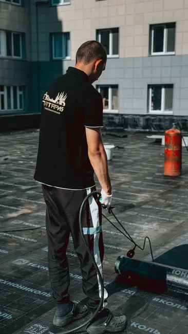 A worker applies bitumen waterproofing on a building roof using a blowtorch for insulation.