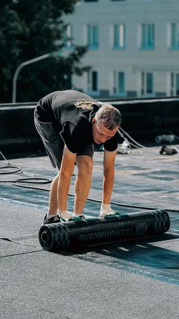 A worker expertly positions a waterproof membrane on a flat roof under clear skies.