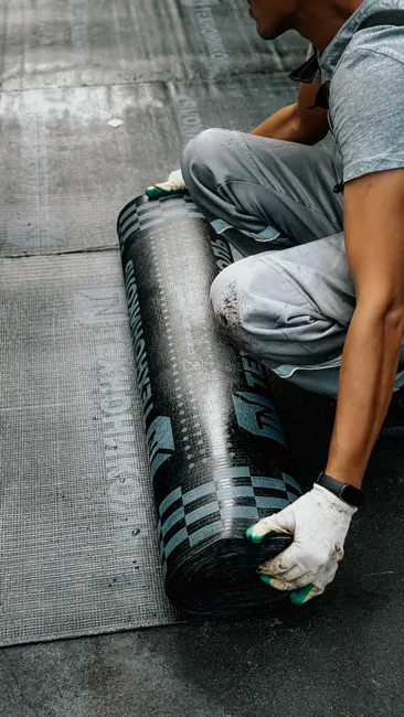 A worker in gloves and helmet installing a waterproof membrane on a flat roof for protection against leaks.