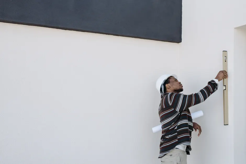 An architect wearing a hard hat checks wall alignment with a level tool outdoors.