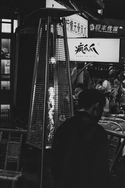Black and white urban scene with a man near a metal grate heater and illuminated signs.