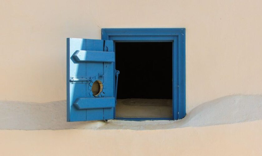 Blue wooden shutter window on a beige wall, open to reveal darkness inside.