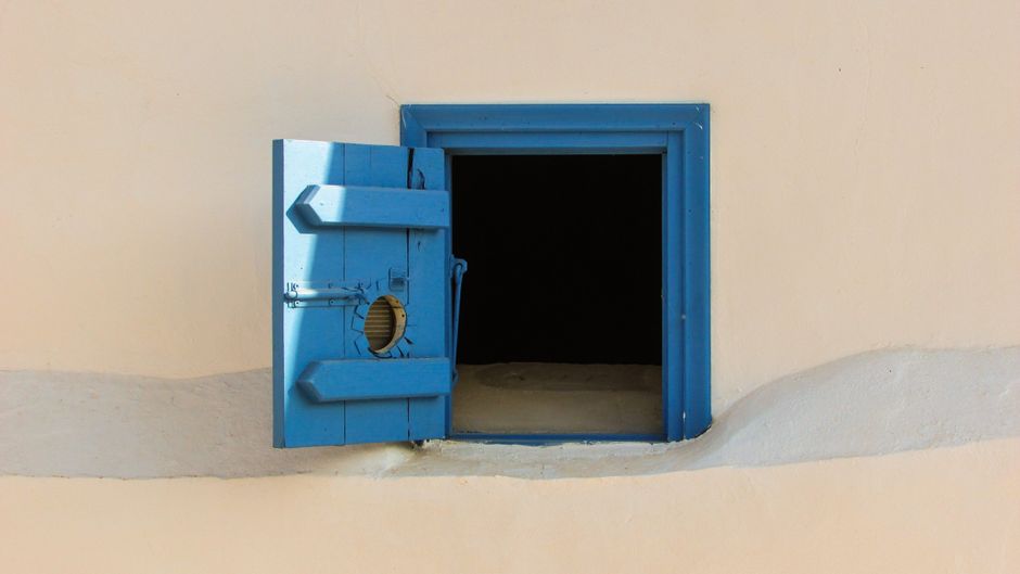 Blue wooden shutter window on a beige wall, open to reveal darkness inside.