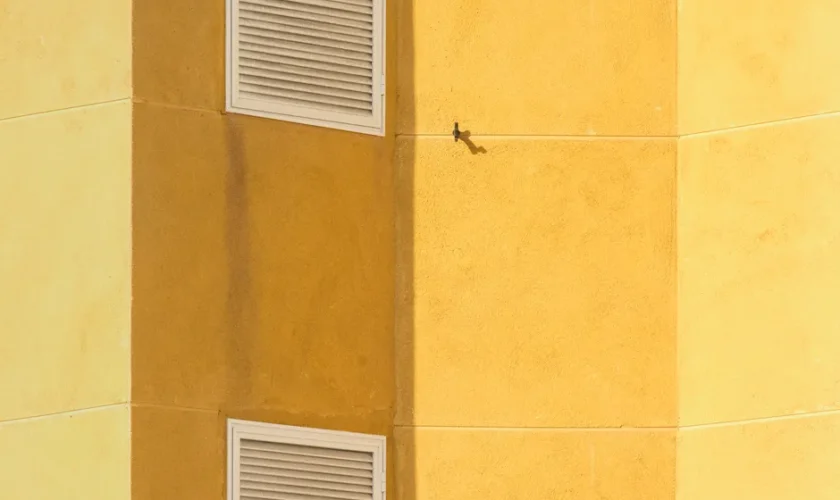 Close-up of a modern yellow building corner featuring ventilation grilles for architectural interest.