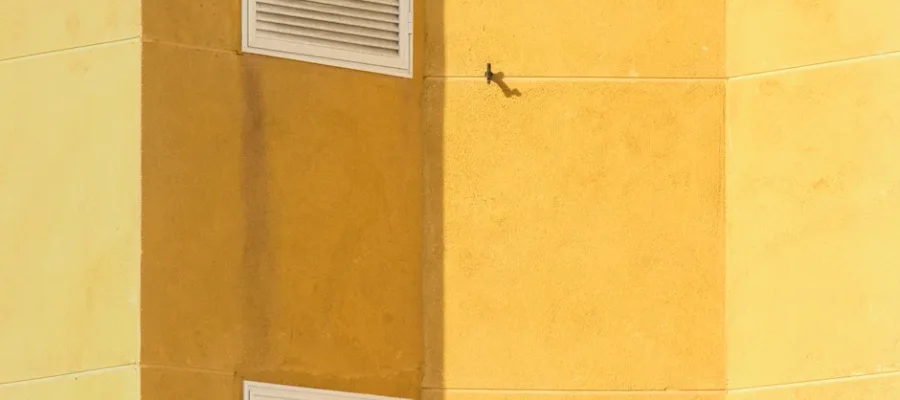 Close-up of a modern yellow building corner featuring ventilation grilles for architectural interest.