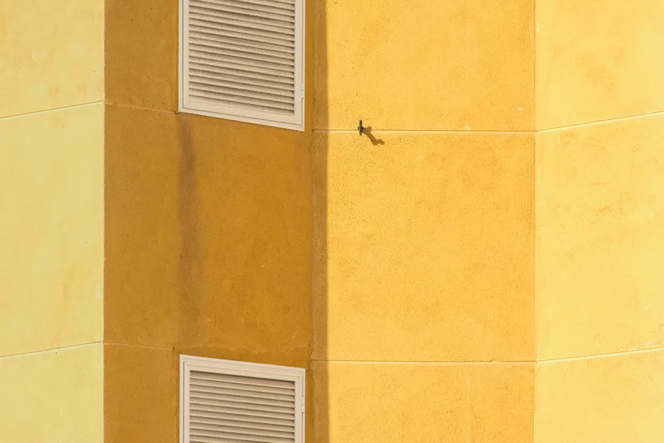 Close-up of a modern yellow building corner featuring ventilation grilles for architectural interest.