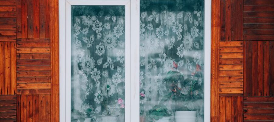 Close-up of a wooden house exterior with a floral curtain window revealing potted plants.