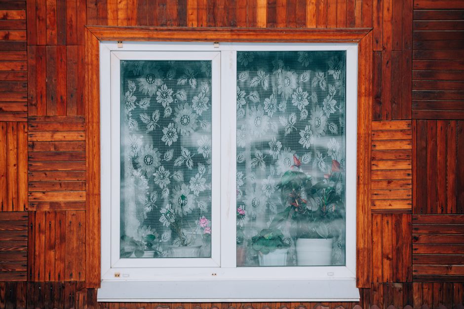 Close-up of a wooden house exterior with a floral curtain window revealing potted plants.