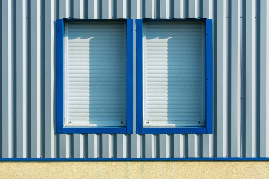 Close-up of corrugated metal wall with blue-framed windows and shutters.
