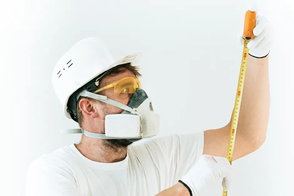 Construction worker using protective gear while measuring with a tape measure indoors.