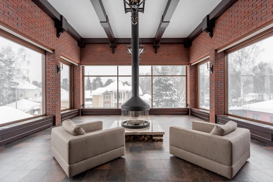 Contemporary living room with armchairs and a central fireplace, snowy view outside.