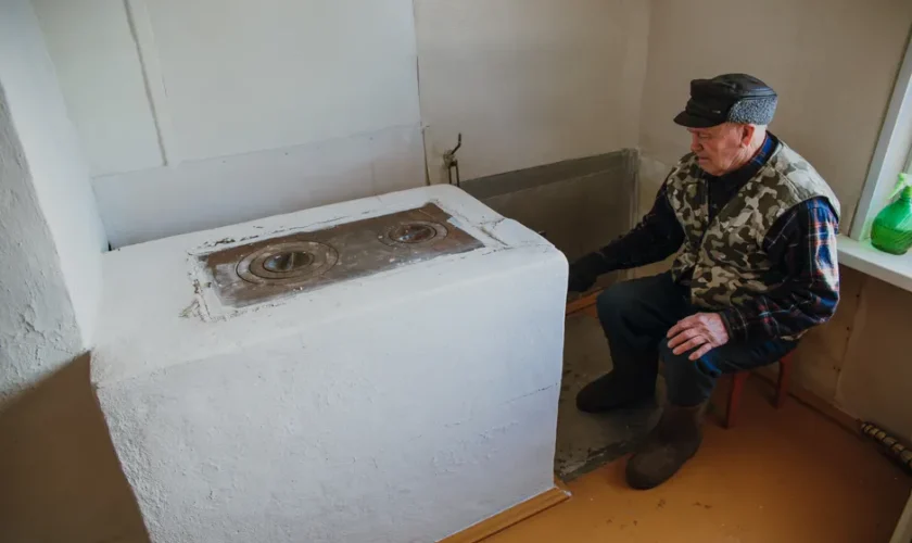 Elderly man wearing warm clothes, sitting by a vintage stove inside a cozy room.