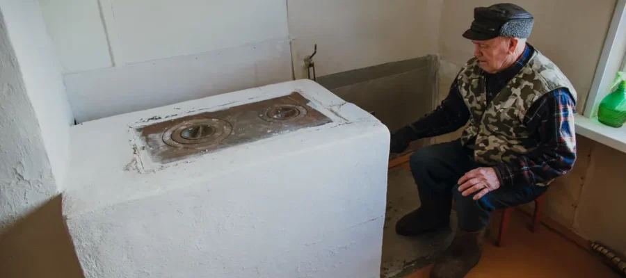 Elderly man wearing warm clothes, sitting by a vintage stove inside a cozy room.