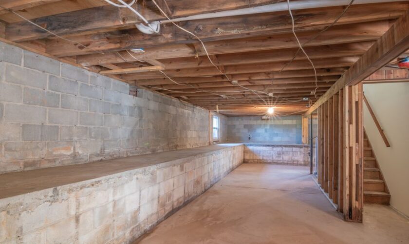 Empty basement with exposed wooden beams, concrete walls and stairs.
