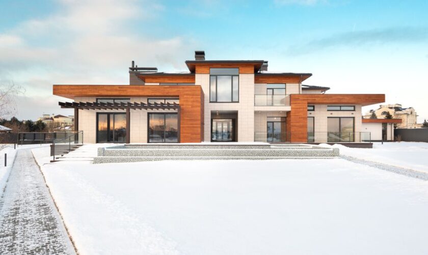 Exterior design of modern cottage house facade with large windows and mixed wooden and stone cladding and spacious yard in snowy winter suburban area