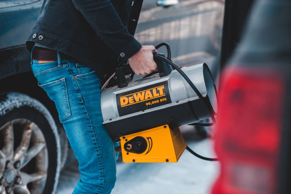 From above side view of crop unrecognizable slim male in jeans carrying portable blow heater with plastic handle and shiny metal tank standing near autos with frozen wheels