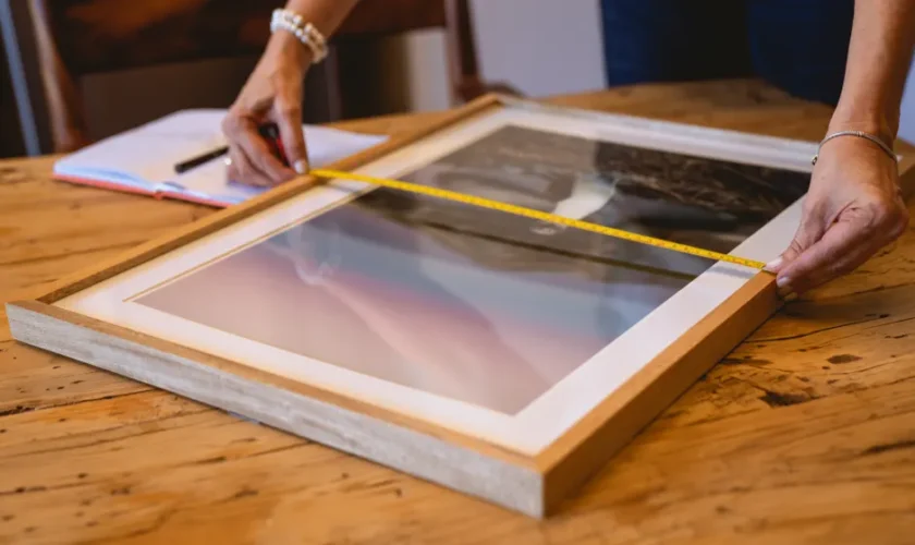 Hands using a tape measure to size a wooden picture frame on a wooden table indoors.