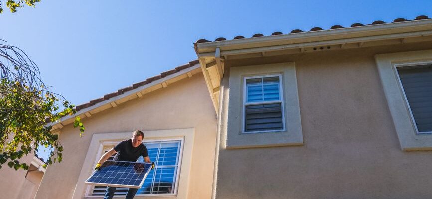 Man installing a solar panel on a rooftop under a clear blue sky, promoting renewable energy.