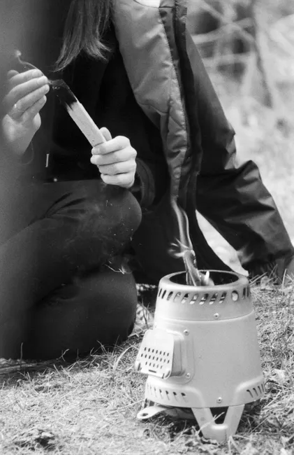 Person sitting by portable stove, preparing a stick for fire, in a black and white photo.