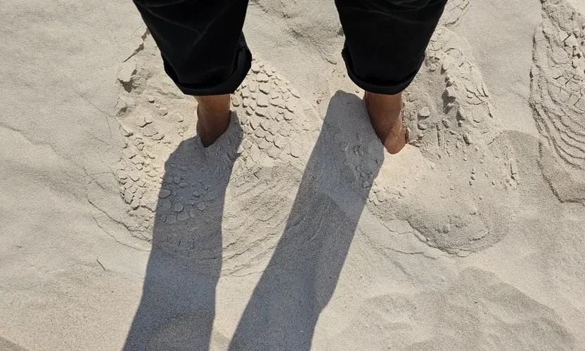 Person with feet buried in sand casting shadows on a sunny beach day.