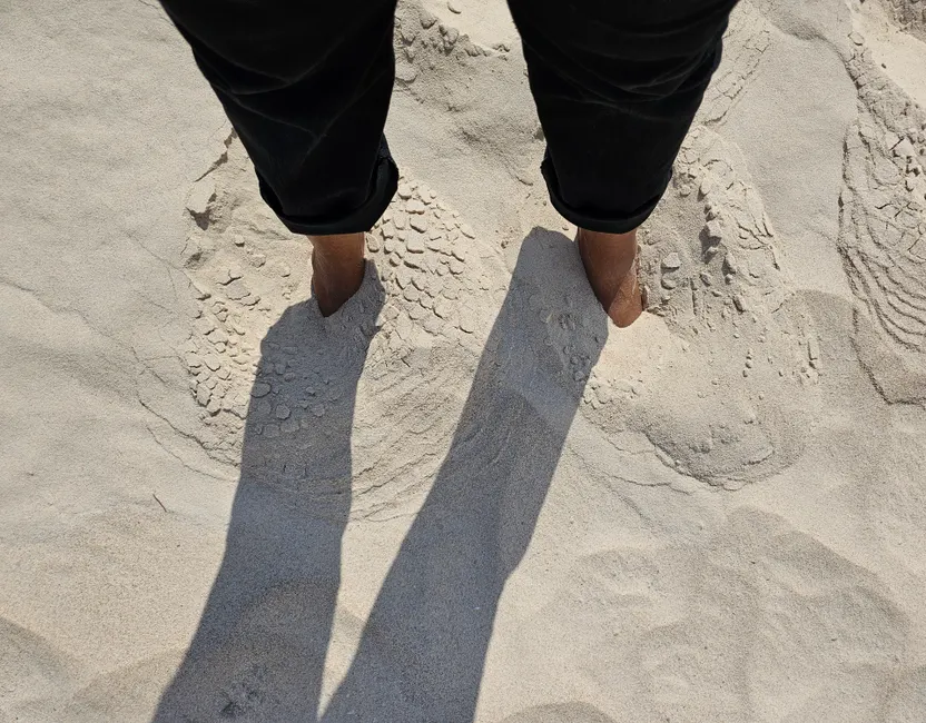 Person with feet buried in sand casting shadows on a sunny beach day.
