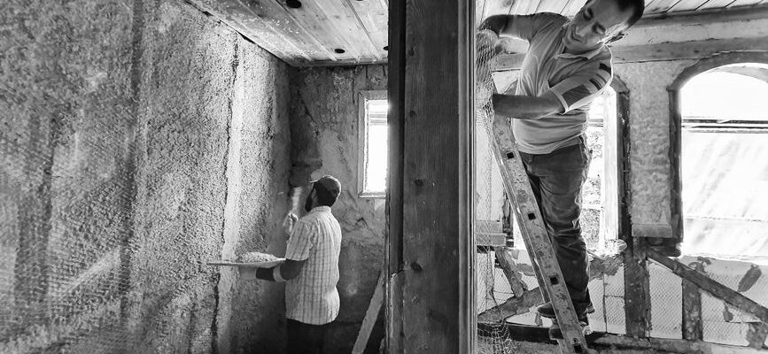 Two men renovating a room, one on a ladder applying plaster, showcasing teamwork and effort.