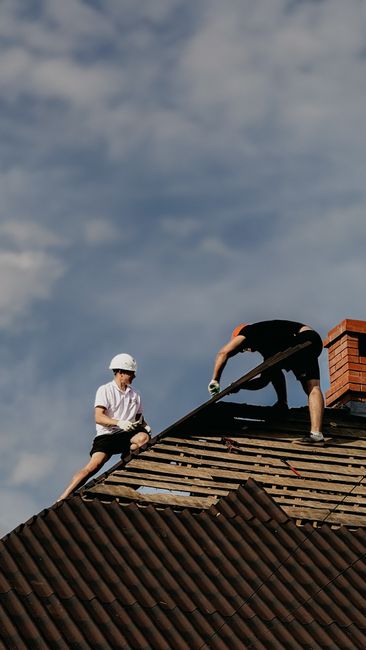 Two roofers working on a roof repair with a clear blue sky, demonstrating teamwork and construction skills.