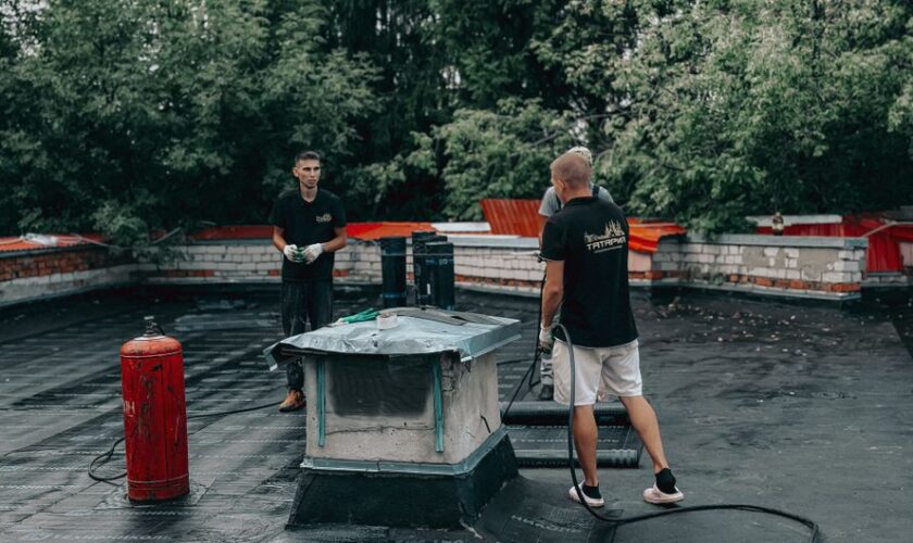 Two workers are performing maintenance on a flat roof with equipment and tools.