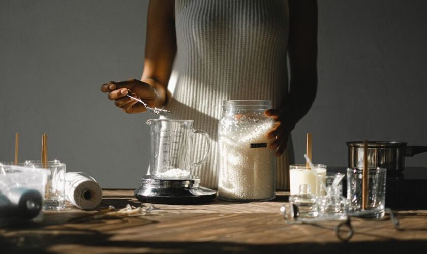 Unrecognizable skilled African American female standing at wooden table a nd weighting wax pellets in jar on scales while making candles