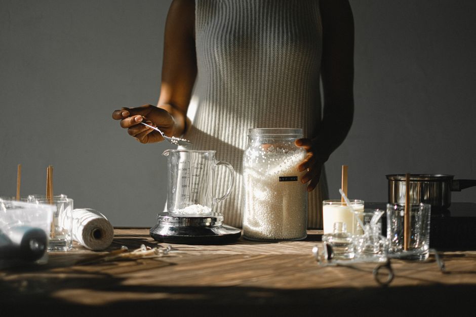 Unrecognizable skilled African American female standing at wooden table a nd weighting wax pellets in jar on scales while making candles