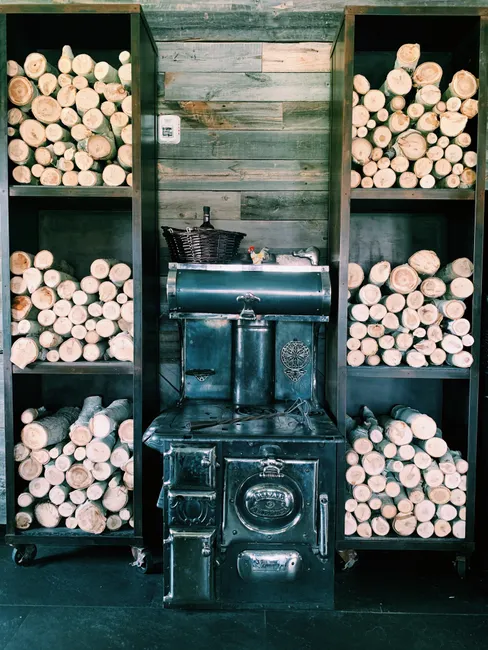 Vintage stove surrounded by shelves of neatly stacked firewood in a rustic indoor setting.