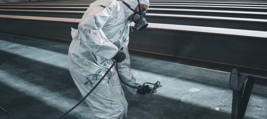 Worker in protective gear spray painting metal beams inside a factory setting.